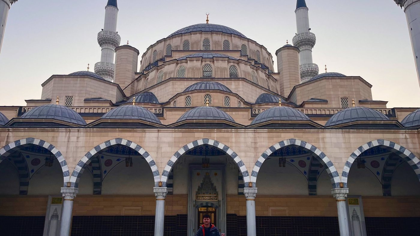 The Central Mosque in Bishkek at dawn — domes, twin minarets and a colonnade of arches, with a visitor standing in the empty courtyard for scale.