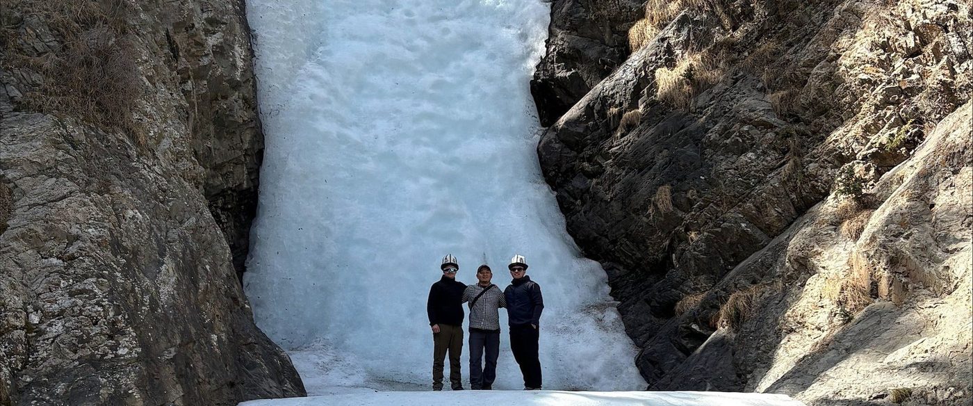 Three men in white Kyrgyz kalpak hats standing at the foot of a frozen waterfall filling a narrow rock gorge.