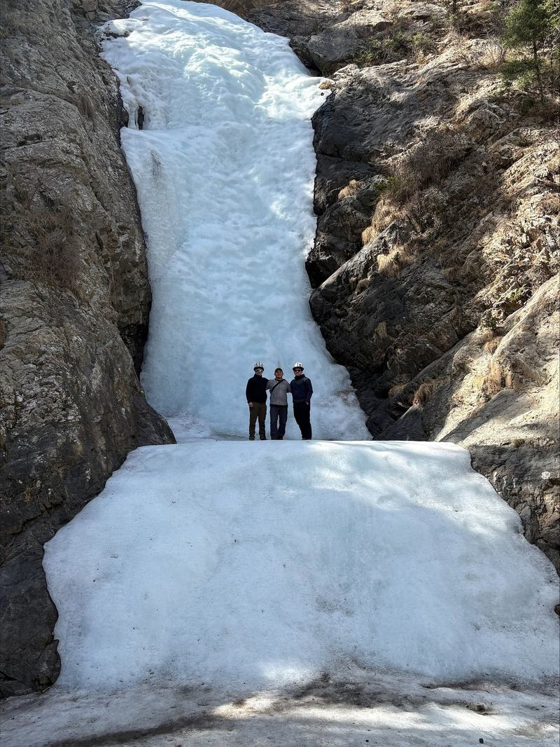 Three men in white Kyrgyz kalpak hats standing at the foot of a frozen waterfall filling a narrow rock gorge.
