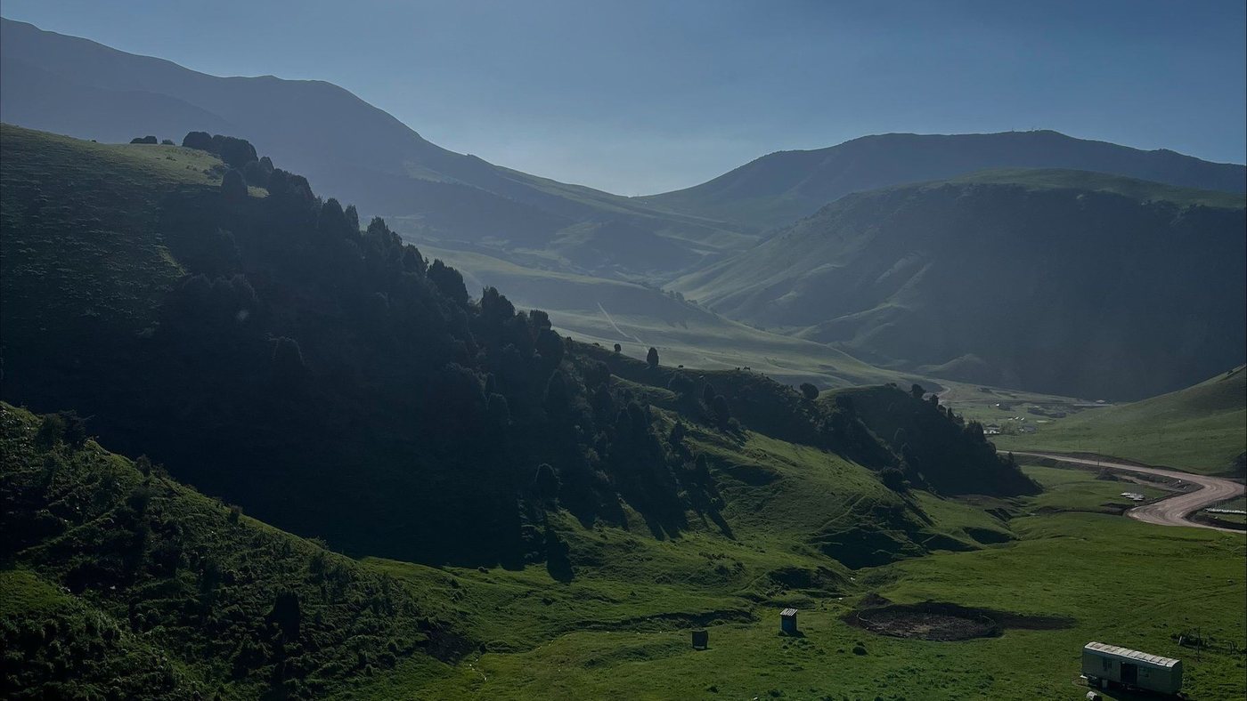 A green summer valley with a stream running through grazing land, forested slopes on one side and mountains beyond, the sun high and flaring.