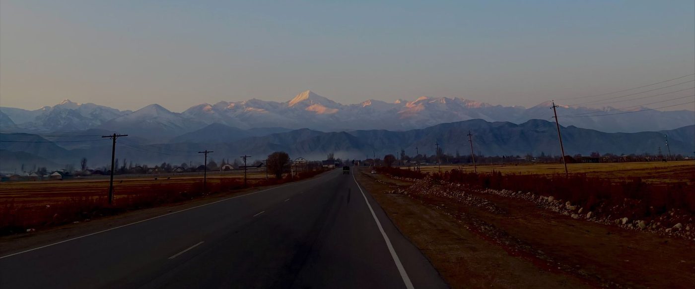 An empty two-lane road running straight towards the snow-covered peaks of the Tian Shan at dusk, with autumn fields and telegraph poles on either side.