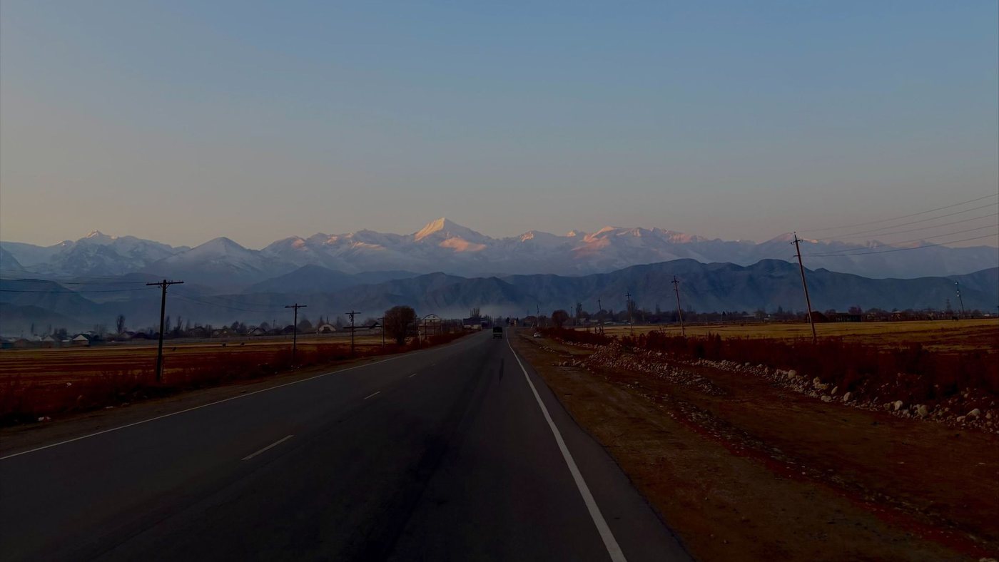 An empty two-lane road running straight towards the snow-covered peaks of the Tian Shan at dusk, with autumn fields and telegraph poles on either side.