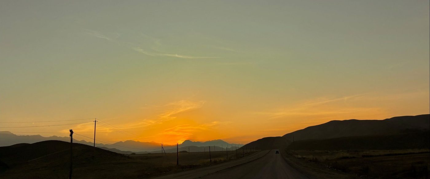 A road running west into an orange sunset over low hills, with a single vehicle in the distance.