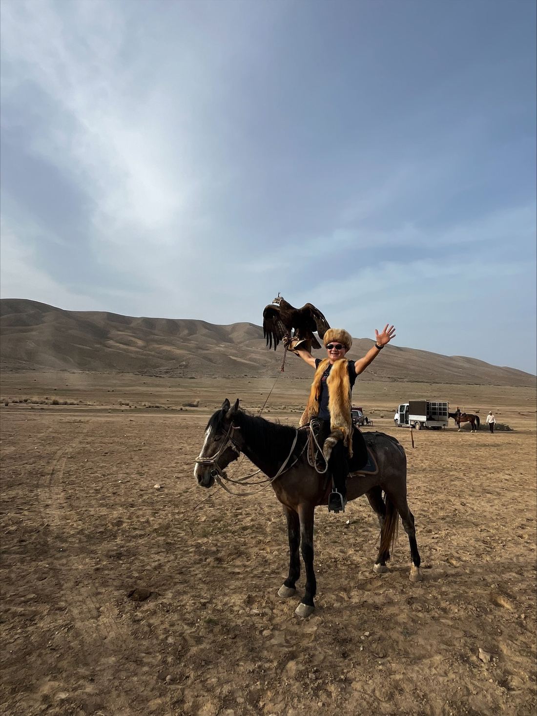 A traveller on horseback in a Kyrgyz kalpak hat holding a golden eagle on a gloved arm, one hand raised, on open ground below dry hills.
